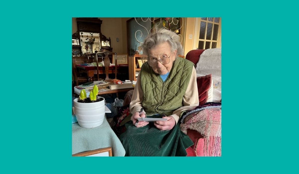Miss Rosemary Tice seated in an arm chair, writing a greetings card, with the Stephen Perse Cambridge birthday hyacinths on the desk beside her.